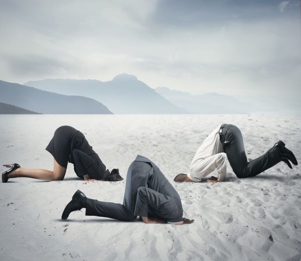Two men and one woman in business attire with their heads in the sand